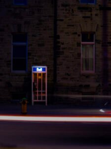 Bell phone booth at night with a large stone wall behind it and a streak of light from a car's lights cut thru the bottom third on the photograph.
